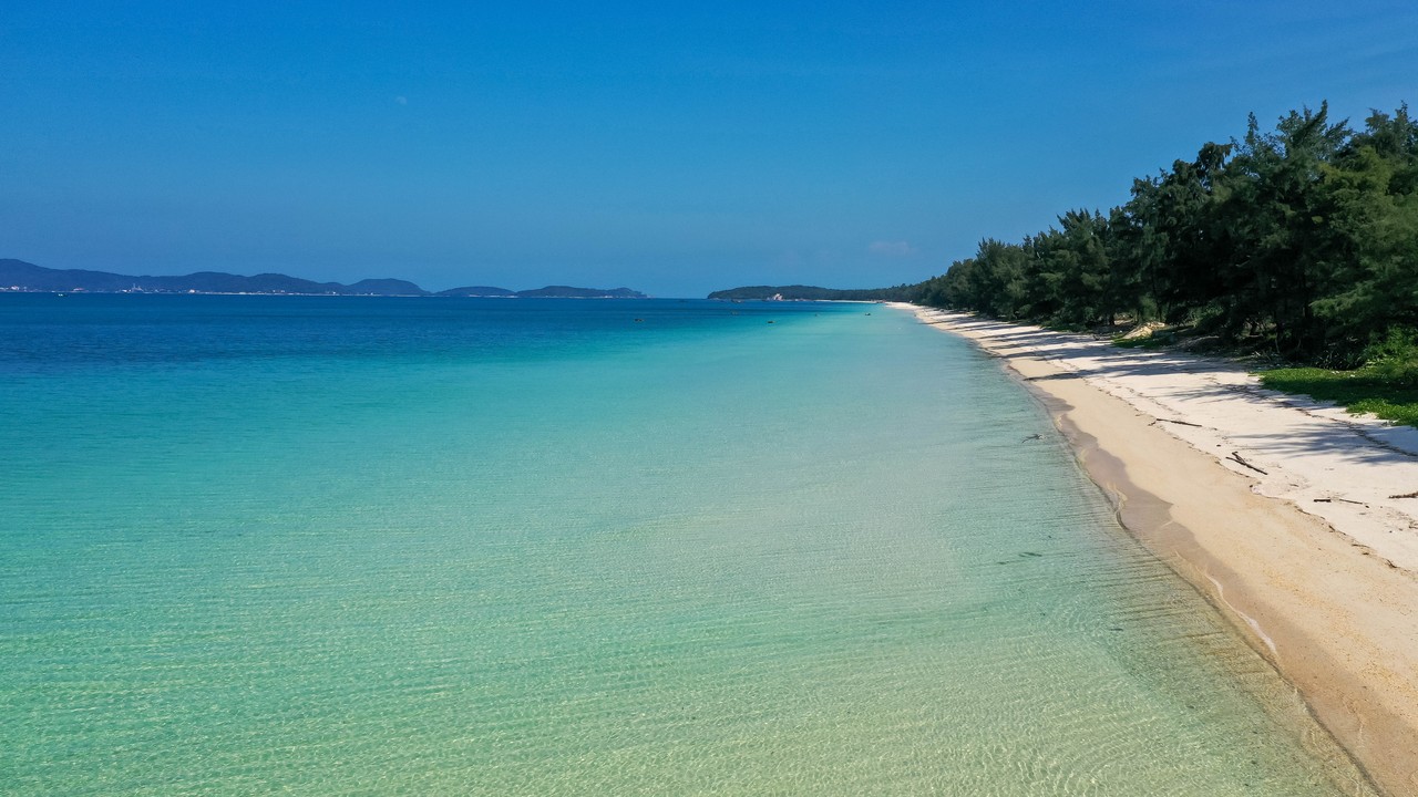 Pale sand and clear shallow water at Hong Van Beach on Co To, with trees lining the coast.