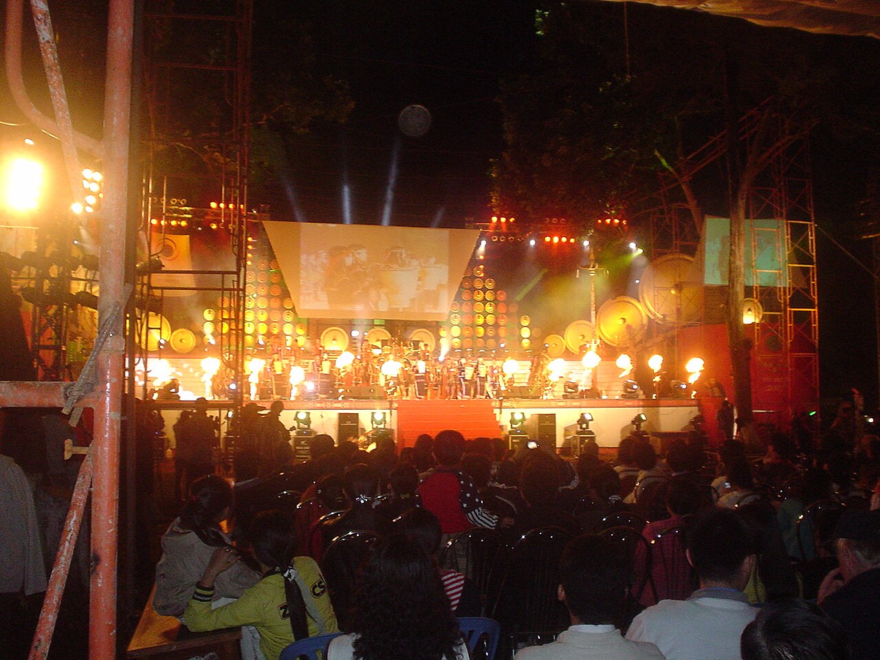 Nighttime gong festival stage in Đắk Lắk, with a semicircle of gongs behind performers and the audience in the foreground; an archival scene.