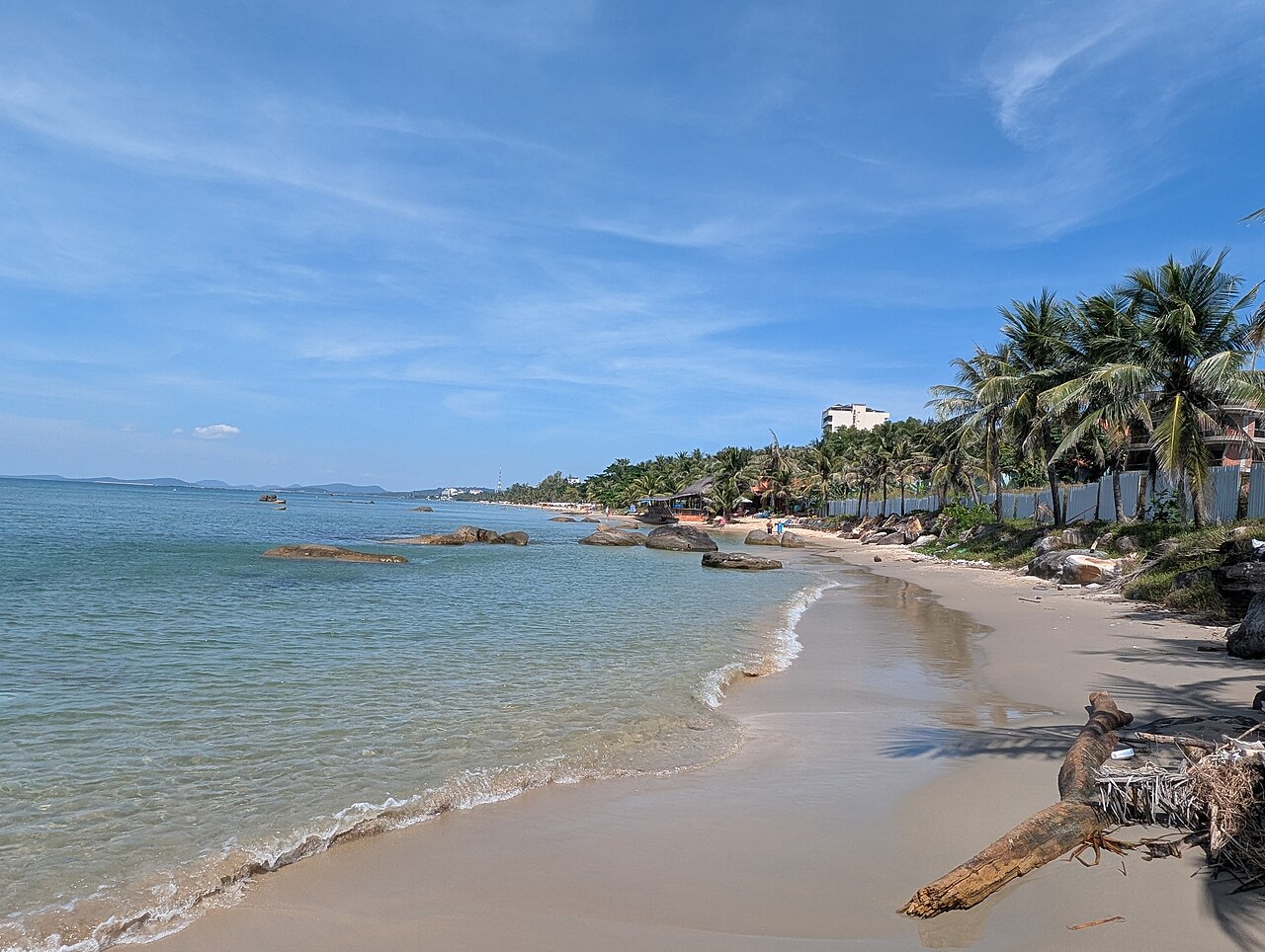 Phú Quốc beach curving past palm trees and low seaside buildings under a bright blue sky.